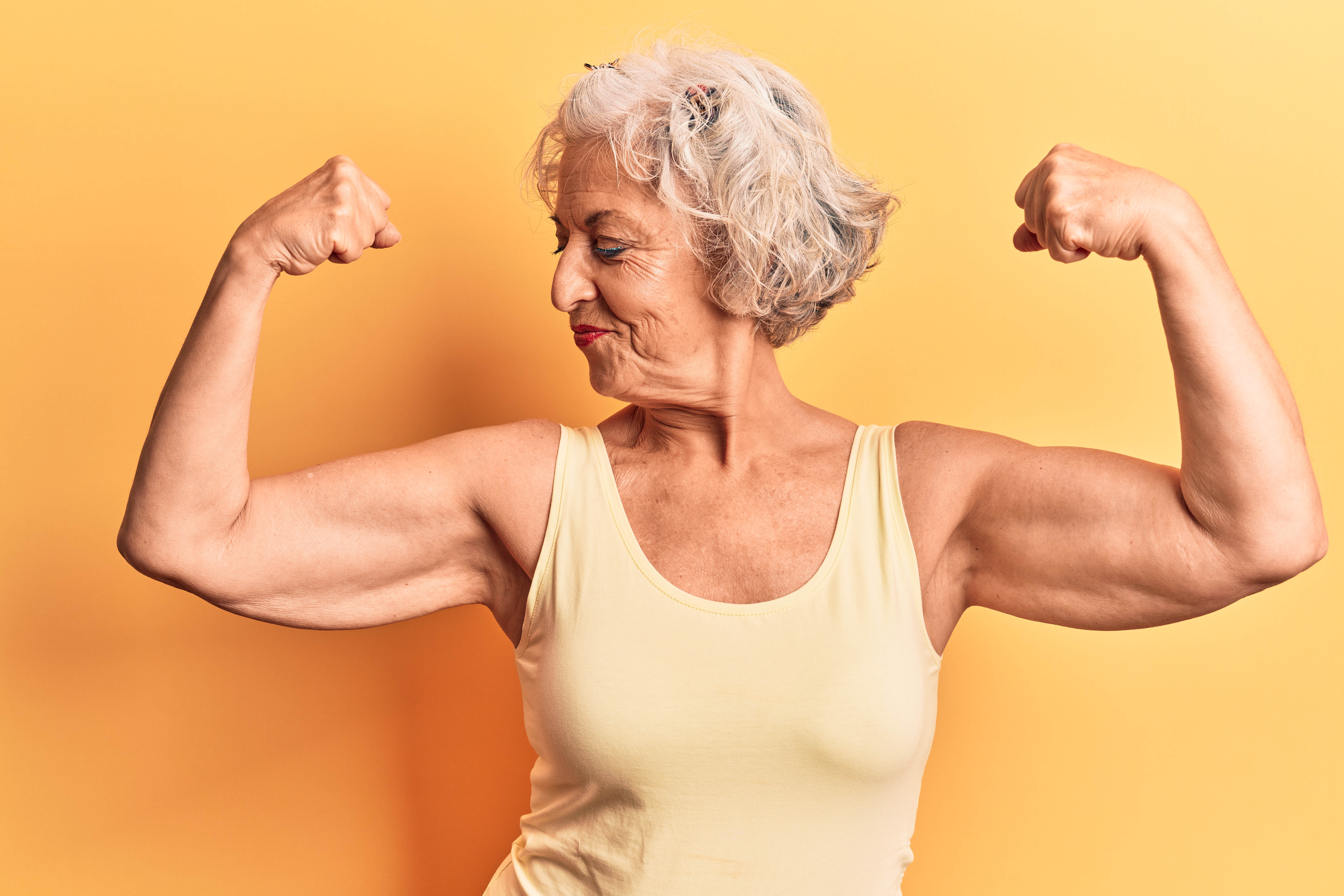 Older woman flexing and looking sideways at her bicep on a yellow background Older woman flexing and looking sideways at her bicep on a yellow background